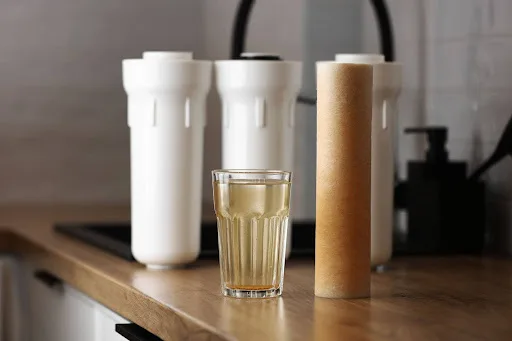 The image shows a close-up of a kitchen counter with two white water filter housings in the background and a used, dirty water filter cartridge standing upright next to a glass of water. The water in the glass has a rusty, brownish coloration, suggesting contamination, which is likely due to the saturated filter cartridge indicating the need for a replacement to ensure clean water.