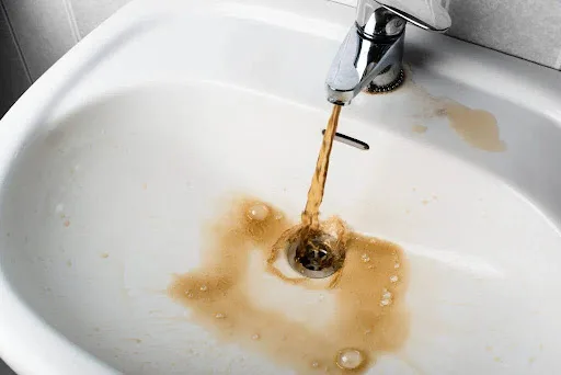 A photo of a sink with water running from the tap showing a high iron content, evidenced by the orange-brown color of the water and staining on the sink.